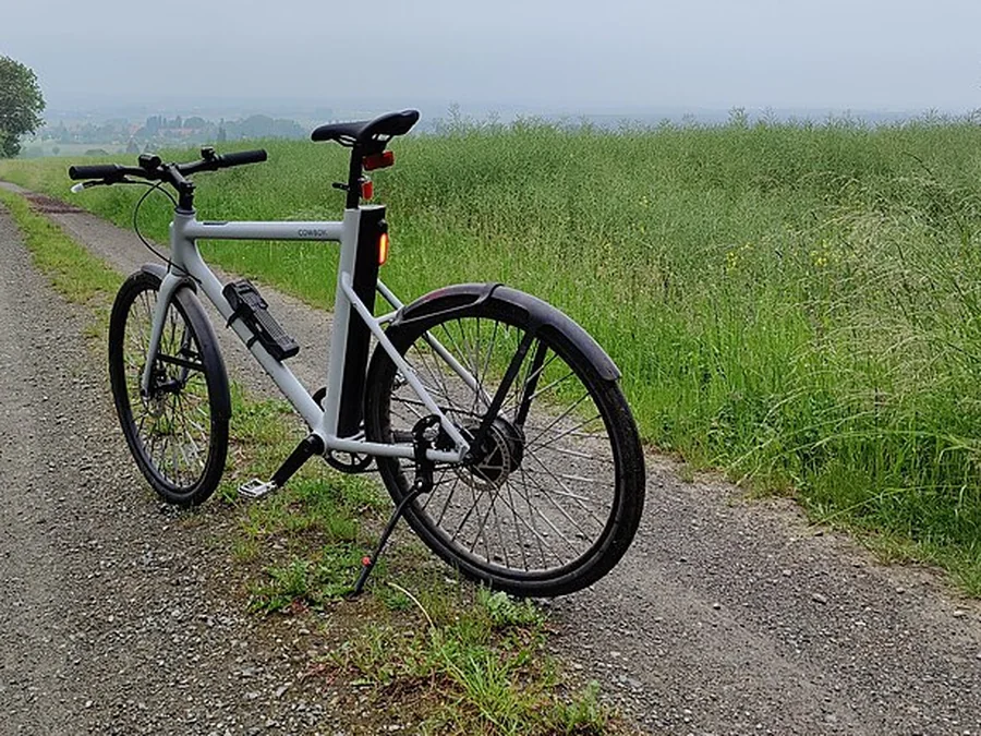 A gray electric bicycle on its kickstand beside a gravel path, with pedals, a saddle and a hub motor visible.