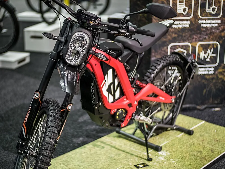 A red Sur-Ron electric dirt bike on a display stand, with footpegs where a bicycle would have pedals.