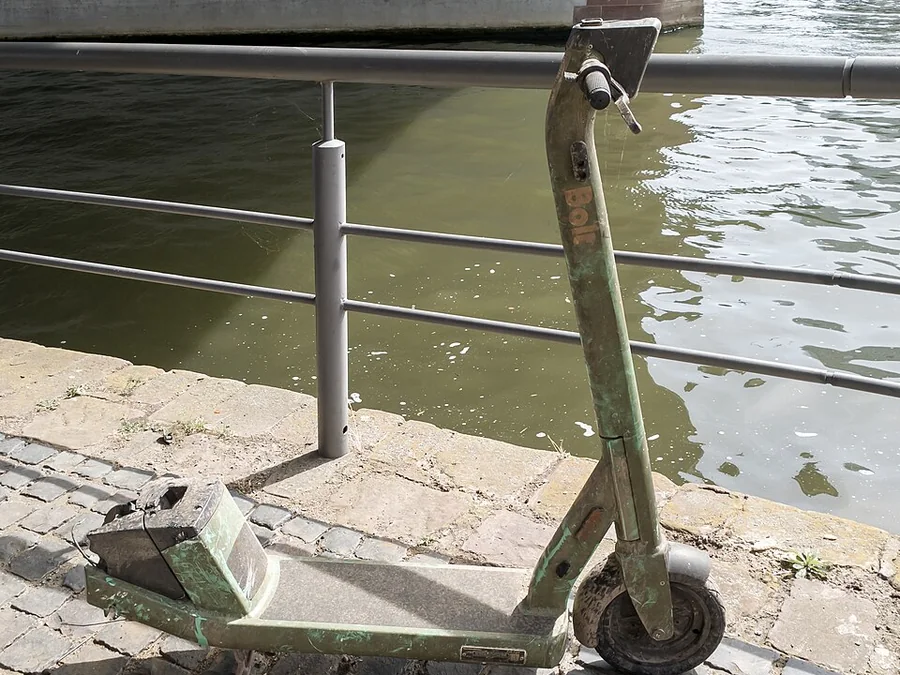 A shared stand-up electric scooter parked upright on a paved riverside walkway, deck and handlebar in view.