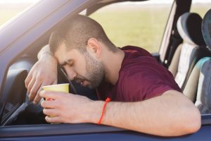 Tired unshaven male leans on wheel and holds paper cup of coffee, has sleepy expression, sits in car, slumbers in automobile, being fatigue to drive all night. Overworked tired young driver.